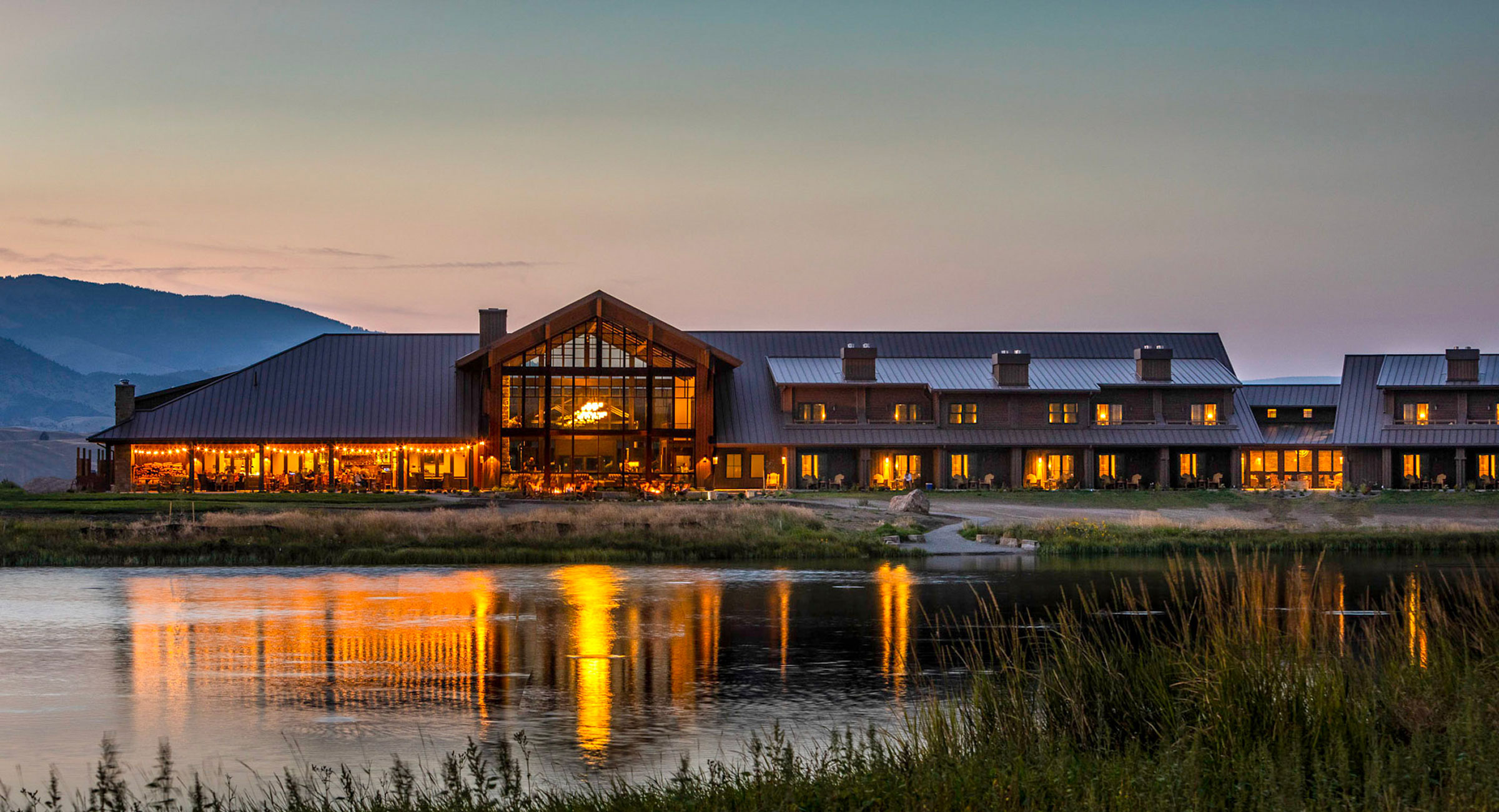 sage-lodge-montana - EXP Journeys - EXP Journeys Lakeside lodge at sunset with warm interior lights reflecting on the water and mountains in the distance **(informative image).**