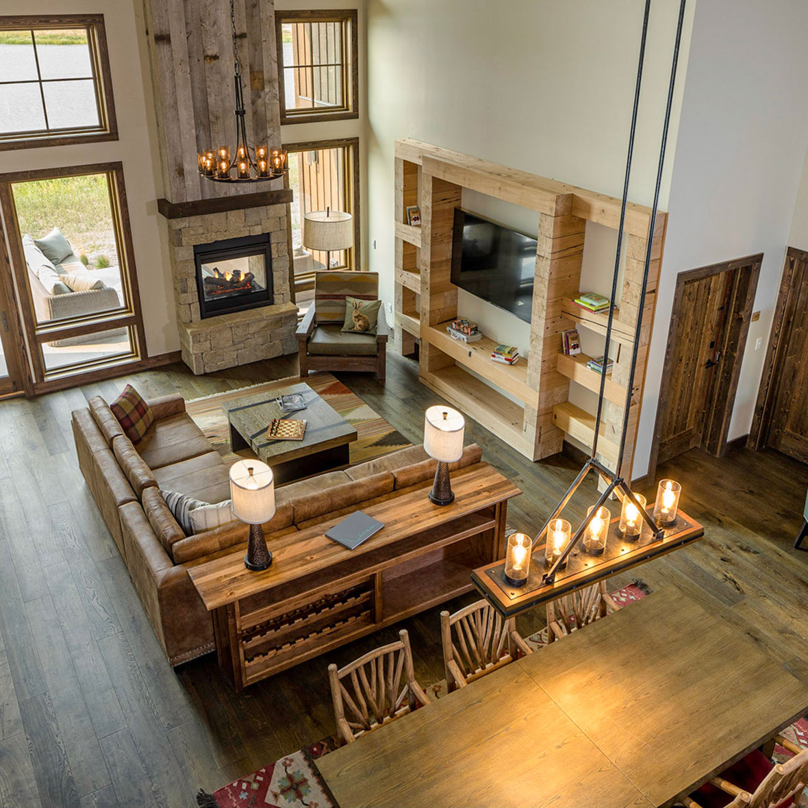sage-lodge-ranch-house - EXP Journeys - EXP Journeys Aerial view of a rustic living room with a stone fireplace, tall windows, and a brown sectional sofa around a wooden coffee table.