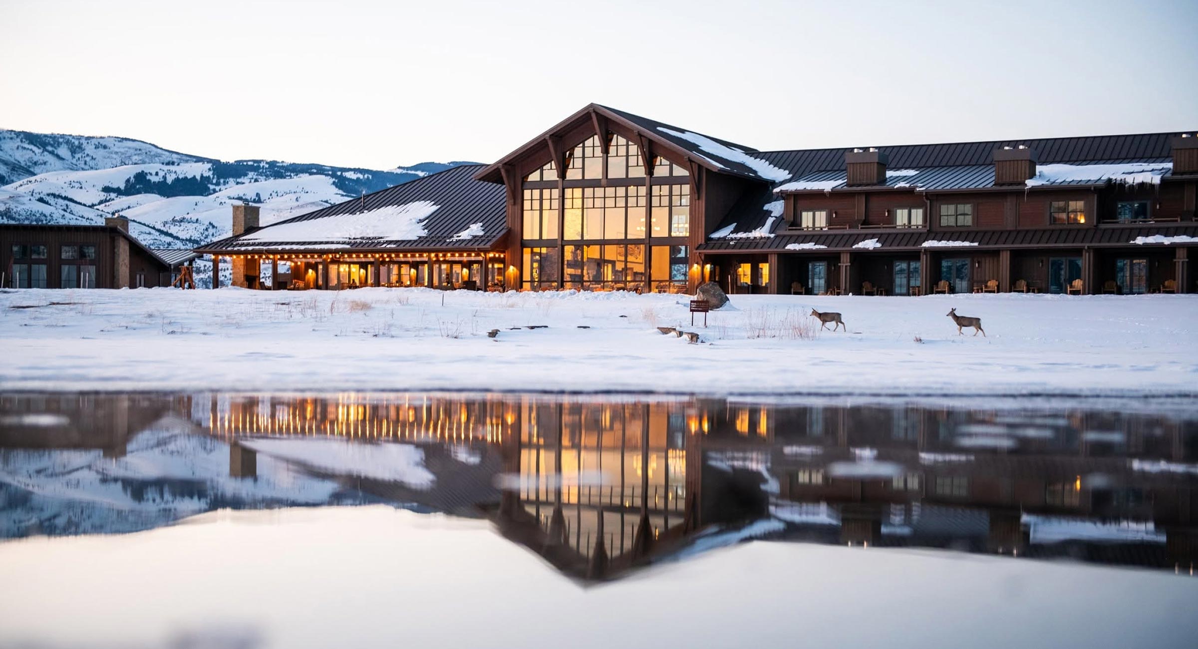 Snowy mountain lodge with lit windows, warm glow along the veranda; snowy hills in the background and two deer near the foreground, reflected in a calm pool.